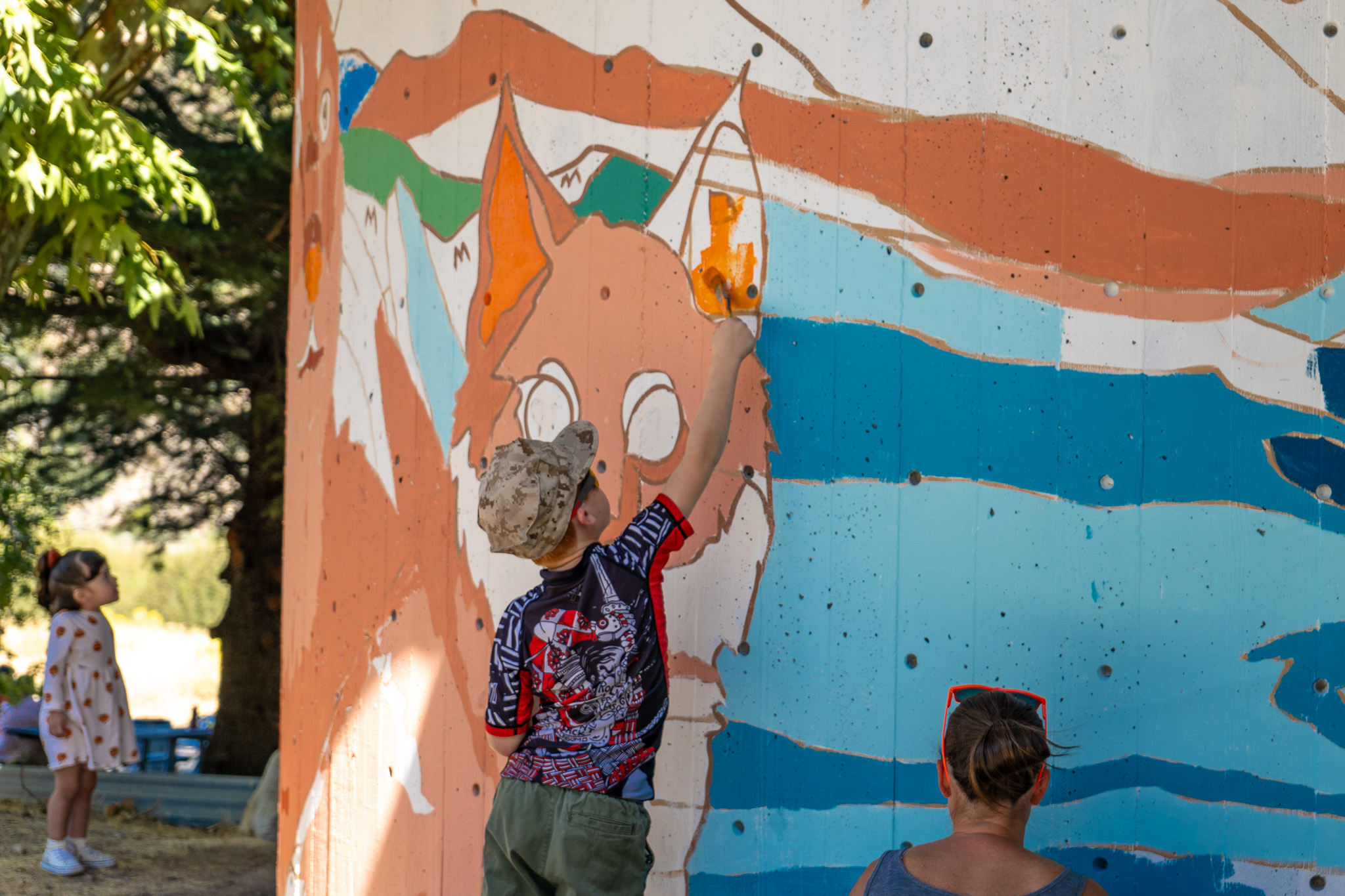 A child reaches up with a paintbrush to color in a stylized bobcat illustration on a large outdoor wall mural. The wall features a paint-by-number design with blue, orange, and white sections. In the background, a young girl in a floral dress looks up, while an adult with sunglasses on their head sits near the base of the wall.