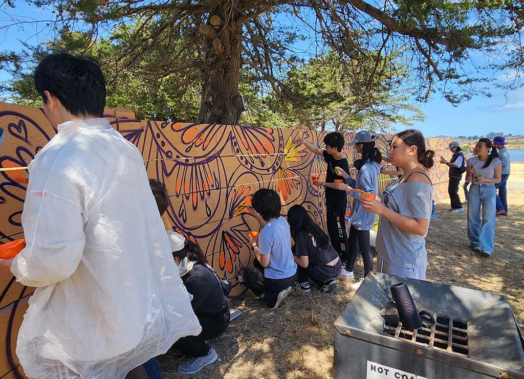 Volunteers of all ages paint a colorful mural on an outdoor wall.