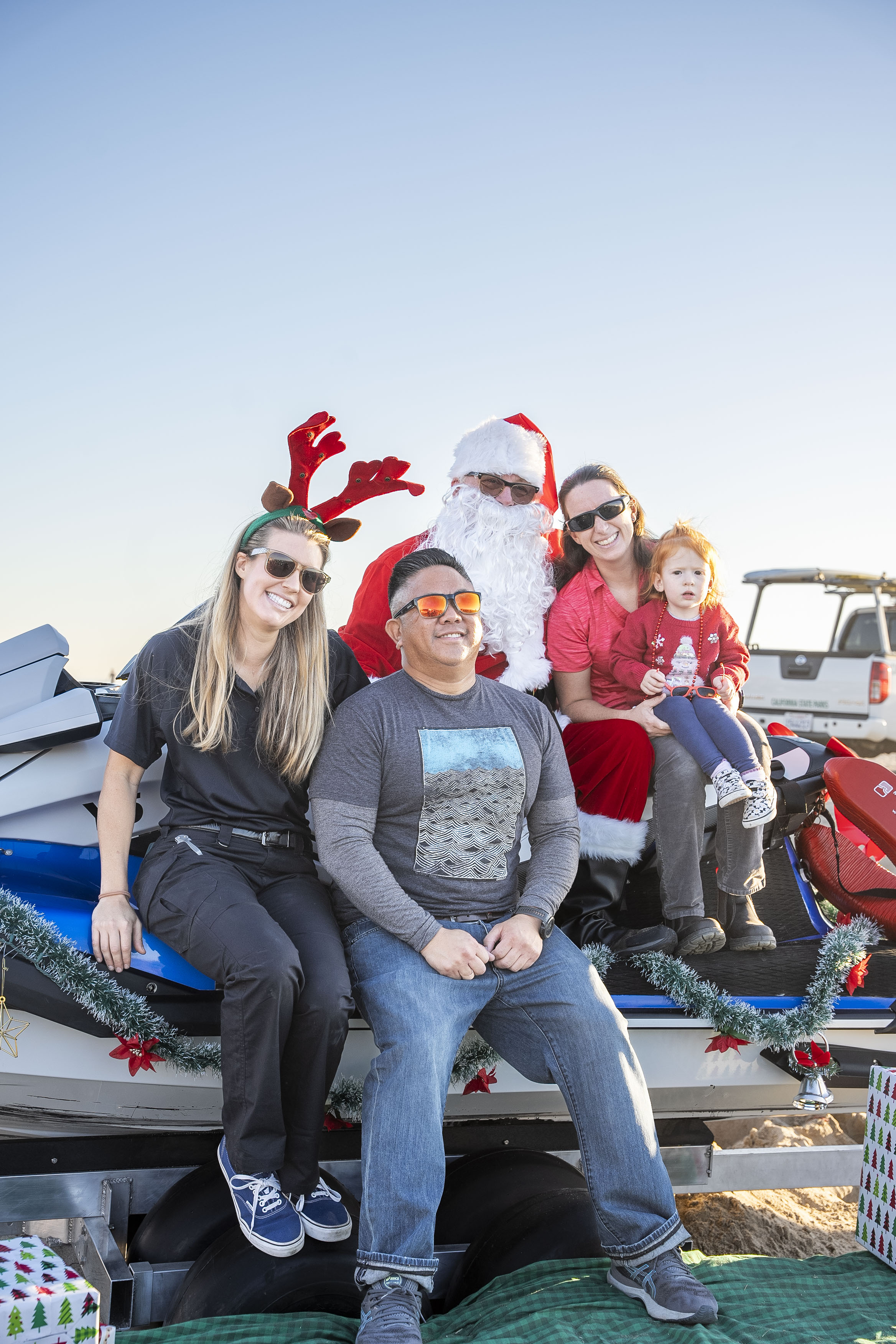 A female lifeguard, a man, and a woman with a young child pose with Santa on his jet ski on the sand at Bolsa Chica.